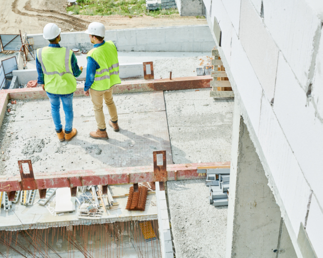 Rear view of contemplative modern constructors in safety vests and hardhats working on new house and discussing building process
